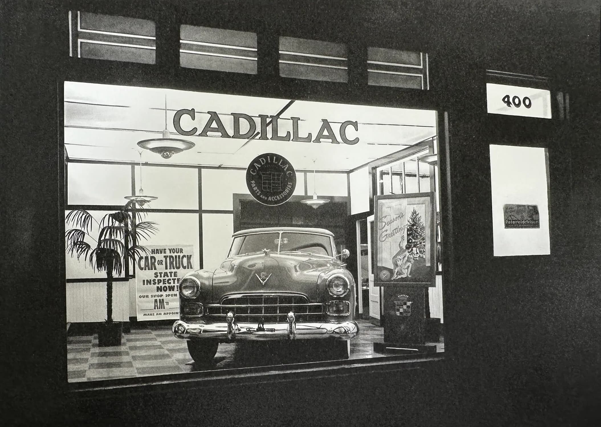 The McGuire Garage showroom lit at night, a new Cadillac behind the plate glass window beneath a CADILLAC sign, with a Season's Greetings display and a potted palm beside it.
